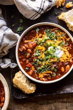 a white bowl filled with chili and beans next to two pieces of bread on a cutting board