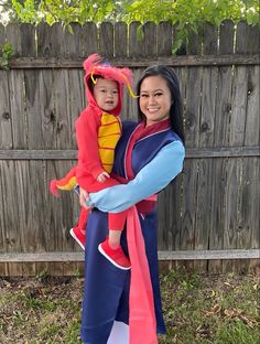 a woman holding a child in her arms while standing next to a fence with a dragon costume on