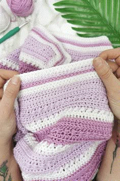 a person holding a crocheted blanket next to a green plant and knitting needles