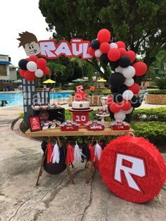 a table topped with balloons and decorations near a swimming pool filled with people in red, white and black