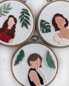 three embroidery hoops with embroidered images of women and leaves on them, hanging from hooks