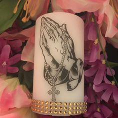 a white candle with a praying hand on it surrounded by pink flowers and gold chains