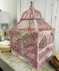 an old pink bird cage sitting on top of a wooden table next to a lamp