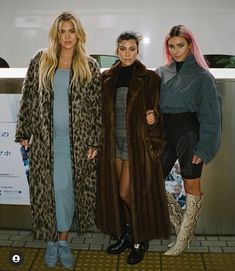 three women standing next to each other in front of a subway station wall wearing long coats and boots