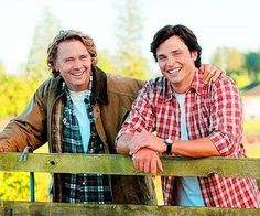 two men standing next to each other near a wooden fence with trees in the background