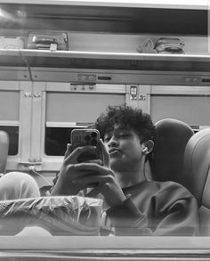 a young man sitting on a train looking at his cell phone and taking a selfie