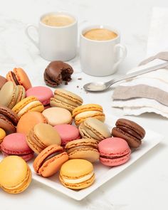 there are many different colored macaroons on the plate next to two cups of coffee