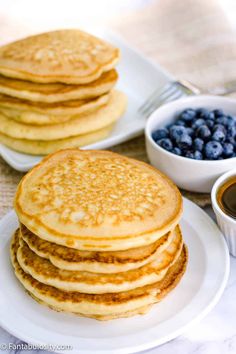 stack of pancakes with syrup and blueberries on white plate next to bowl of fruit