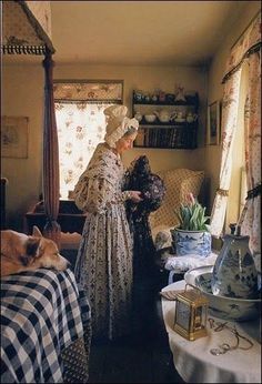 two women in period dress standing next to each other near a table with a potted plant on it