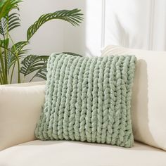 a green knitted pillow sitting on top of a white couch next to a potted plant