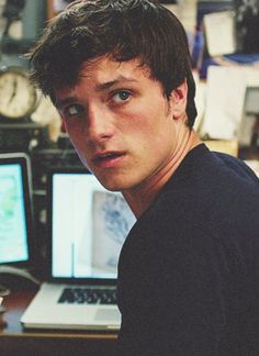 a young man sitting in front of a computer desk with two monitors and a clock on the wall