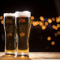 two glasses of beer sitting on top of a table next to each other with blurry lights in the background