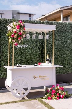 a white cart with flowers on it sitting in front of a wall and some wine glasses