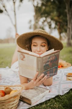 a woman laying on the grass reading a book while wearing a straw hat and holding an apple in her hand