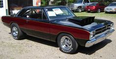 a red and black muscle car parked in front of a garage with other cars behind it