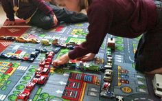 two children playing with toy cars on a play mat in a room full of people