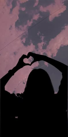 the silhouette of a woman making a heart shape with her hands against a cloudy sky