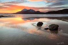 two large rocks sitting on top of a sandy beach next to the ocean at sunset