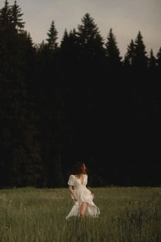 a woman in a white dress is walking through tall grass with trees in the background