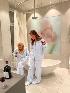 two women standing in front of a bath tub holding wine glasses