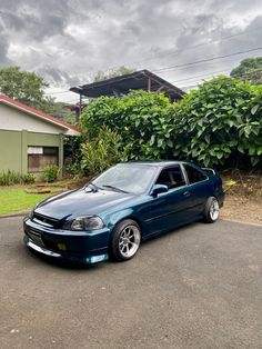 a blue car is parked in front of a house with bushes and trees behind it