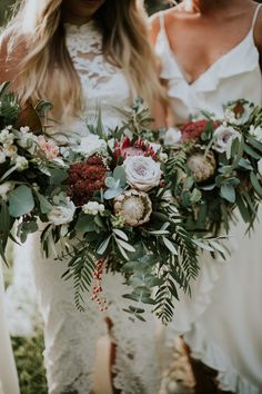 two women in white dresses holding bouquets of flowers and greenery on their hands