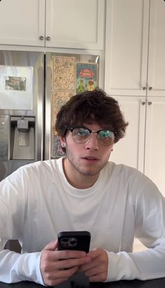 a young man sitting at a kitchen counter looking at his cell phone while wearing glasses