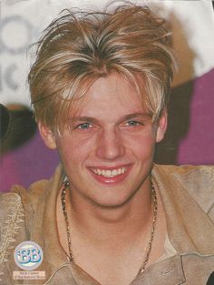 a young man with blonde hair wearing a necklace and leather jacket smiling at the camera