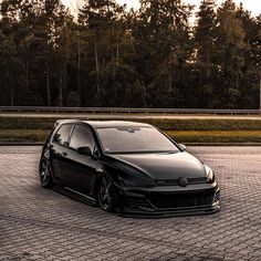 a black car parked on top of a brick parking lot next to some tall trees