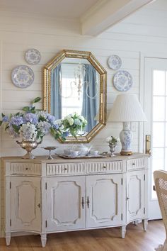 a white dresser with blue and white plates on it next to a large gold framed mirror