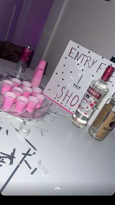 a table topped with lots of pink cups next to a bottle of alcohol and a sign