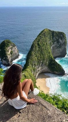 a woman sitting on the edge of a cliff overlooking the ocean and two rock formations