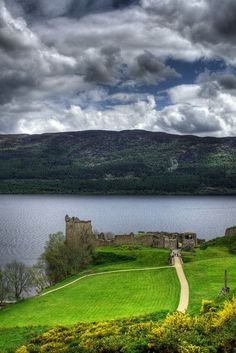 an old castle sitting on top of a lush green hillside next to a lake under a cloudy sky