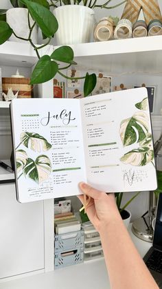 a person holding an open planner in front of a desk with plants and books on it