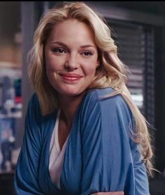 a woman with blonde hair wearing a blue jacket and smiling at the camera in a kitchen