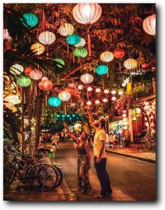 two people standing in the middle of a street with many lanterns hanging from the ceiling