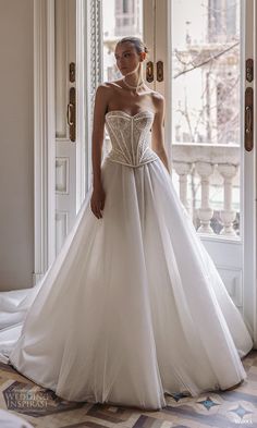 a woman standing in front of a window wearing a wedding dress with a bow on the waist