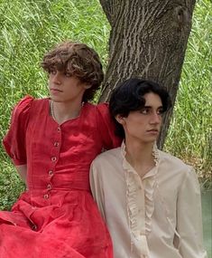 two people sitting next to each other in front of a tree with water behind them