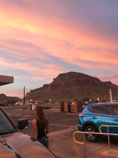 a woman standing next to a car in a parking lot with mountains in the background