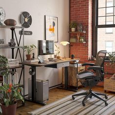 an office with a brick wall and lots of plants on the desk, in front of a large window