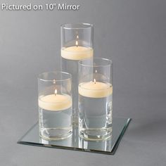 three clear glass vases with lit candles in them sitting on a mirrored tray against a gray background