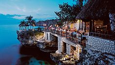 people are sitting at tables on the edge of a cliff overlooking the ocean and trees