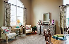 a living room filled with furniture next to two large windows and a wooden dining table