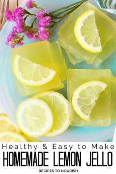 A photo with a clear blue glass plate topped with about seven 2 inch squares of homemade lemon jello, stacked in 3 stacks, with a small sprig of bright pink Statice dried flowers sitting next to it, with two forks and a white and blue waves paisley print kitchen towel sitting under the plate and lemon slices and a half a lemon sitting around the plate with text that says Real Food Homemade Grassfed Gelatin Healthy Lemon Jello.