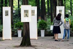 two women standing in front of three framed pictures and trees with potted plants behind them