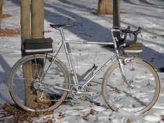 a silver bicycle parked next to a tree in the snow near a pole with a bag on it