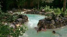 two people are swimming in a pool with rocks and trees around it, surrounded by greenery