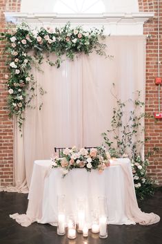 a table with candles and flowers on it in front of a brick wall, surrounded by greenery