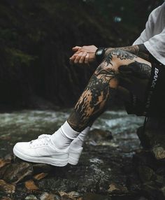 a man with tattoos sitting on top of rocks next to a river wearing white sneakers