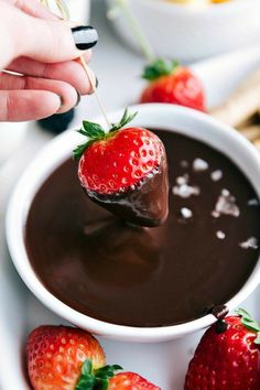 someone dipping strawberries into chocolate sauce in a white bowl with other fruit on the side